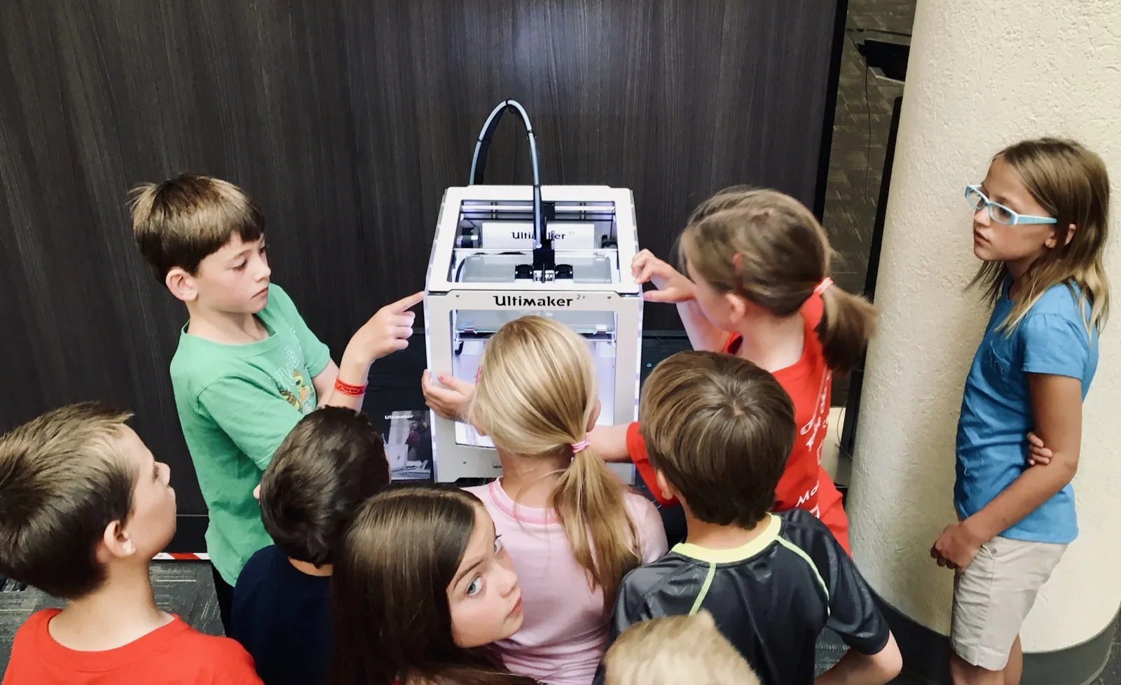A cluster of children gathered around a 3D printer, watching a part come to life