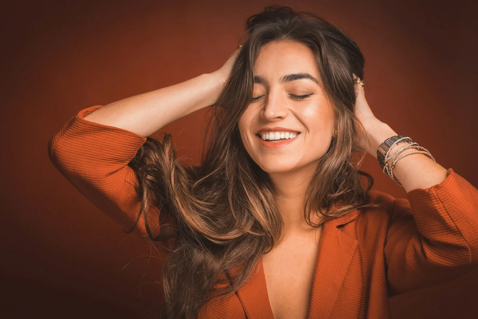 A guest laughing, hands lifted through freshly painted caramel balayage, on a warm rust studio ground.